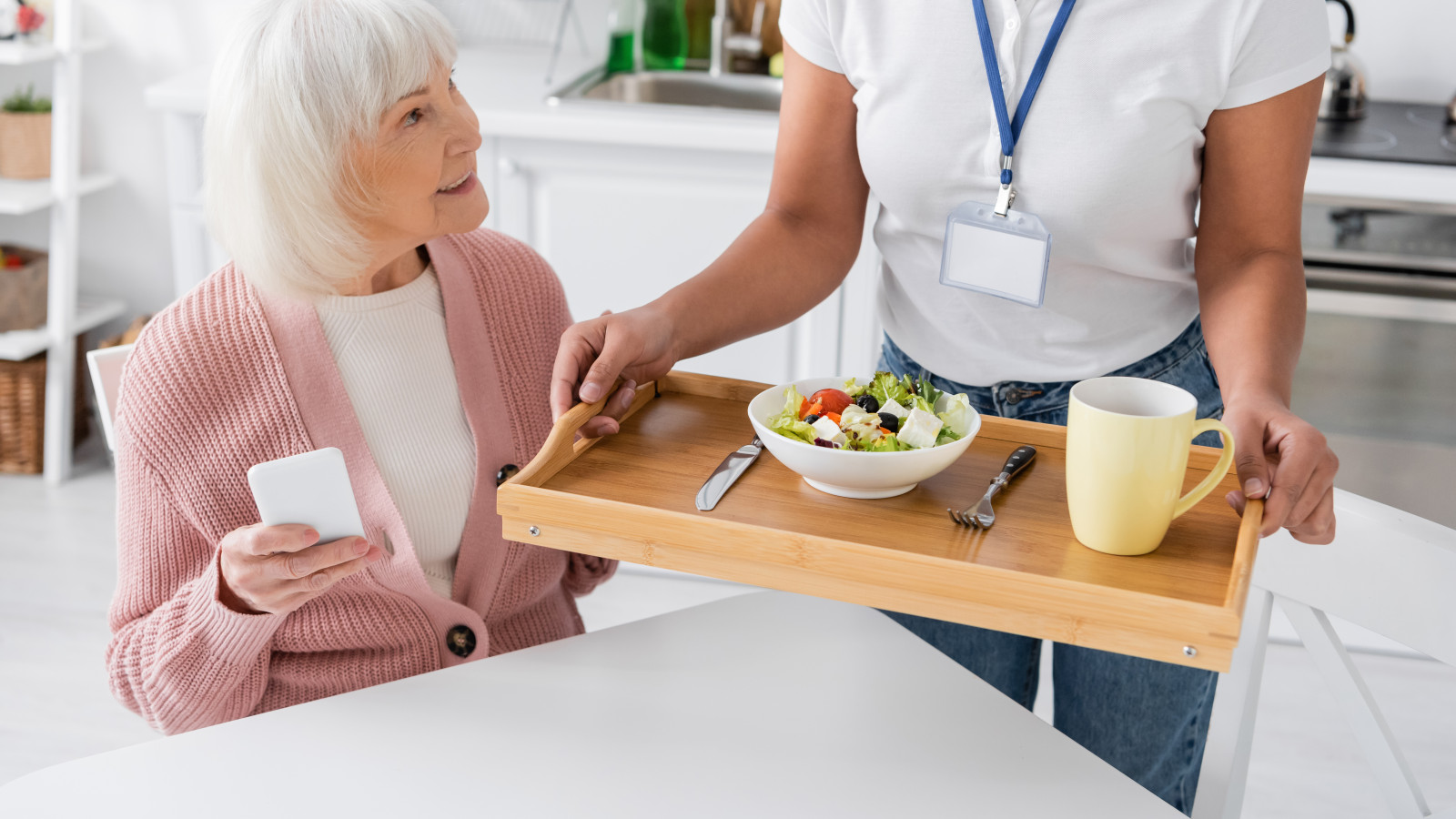 Ältere Frau mit Handy erhält von einer Person ein Tablett mit Essen und Getränk.